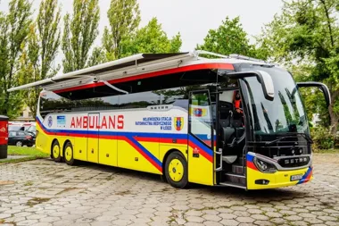 Specjalistyczny autobus ambulansowy Wojewódzkiej Stacji Ratownictwa Medycznego w Łodzi stoi na brukowanym placu wśród drzew.