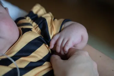 Dłoń niemowlęcia w żółto-czarnej koszulce w pasy trzyma palec dorosłej osoby.
