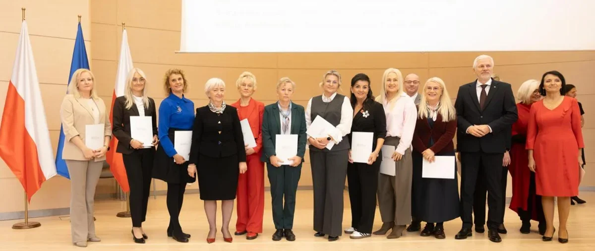 Grupa osób stoi na sali konferencyjnej z flagami w tle podczas uroczystości wręczania dokumentów.
