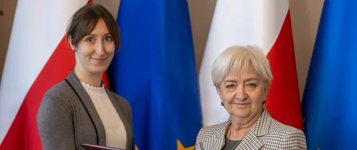 Dwie kobiety pozują do zdjęcia przed flagami Polski i Unii Europejskiej podczas oficjalnej uroczystości.